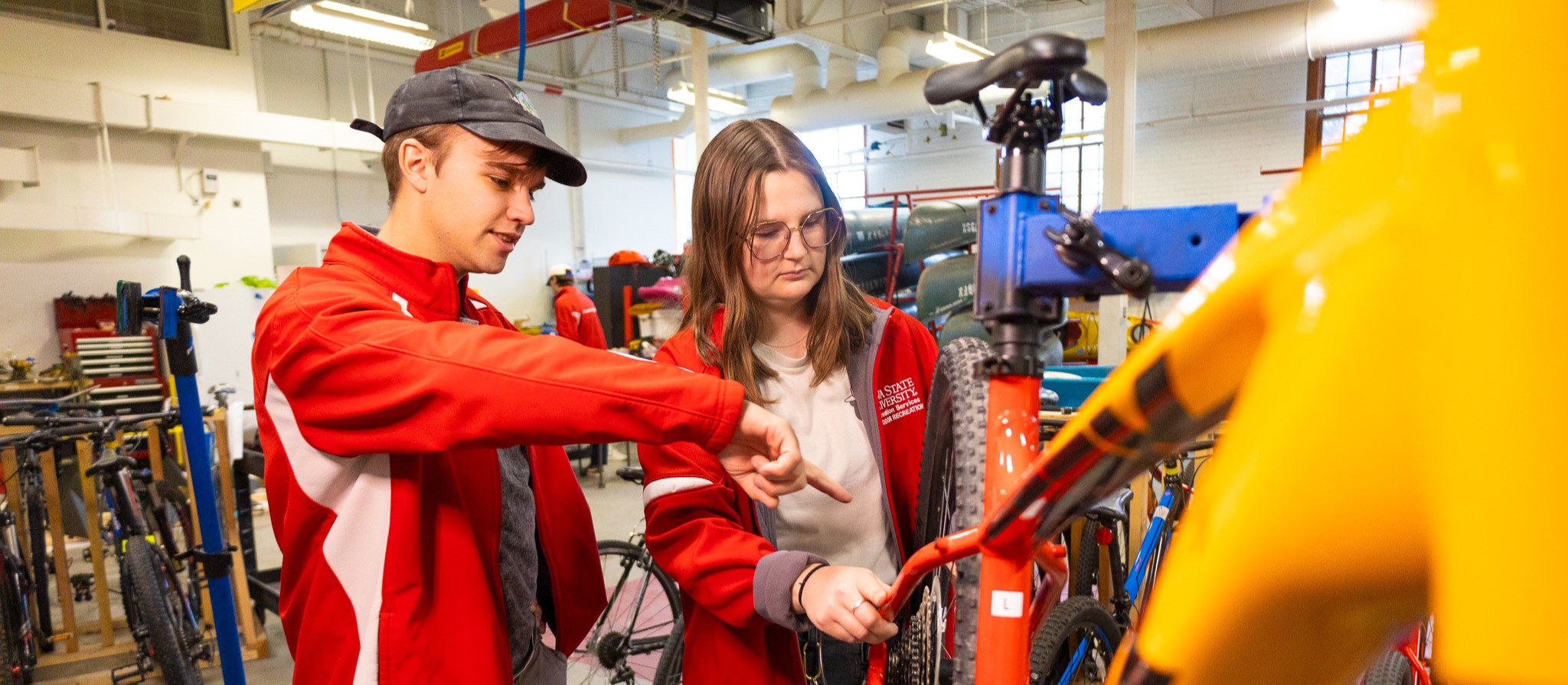 Two college aged individuals, one male and one female, work on repairing a yellow bicycle.