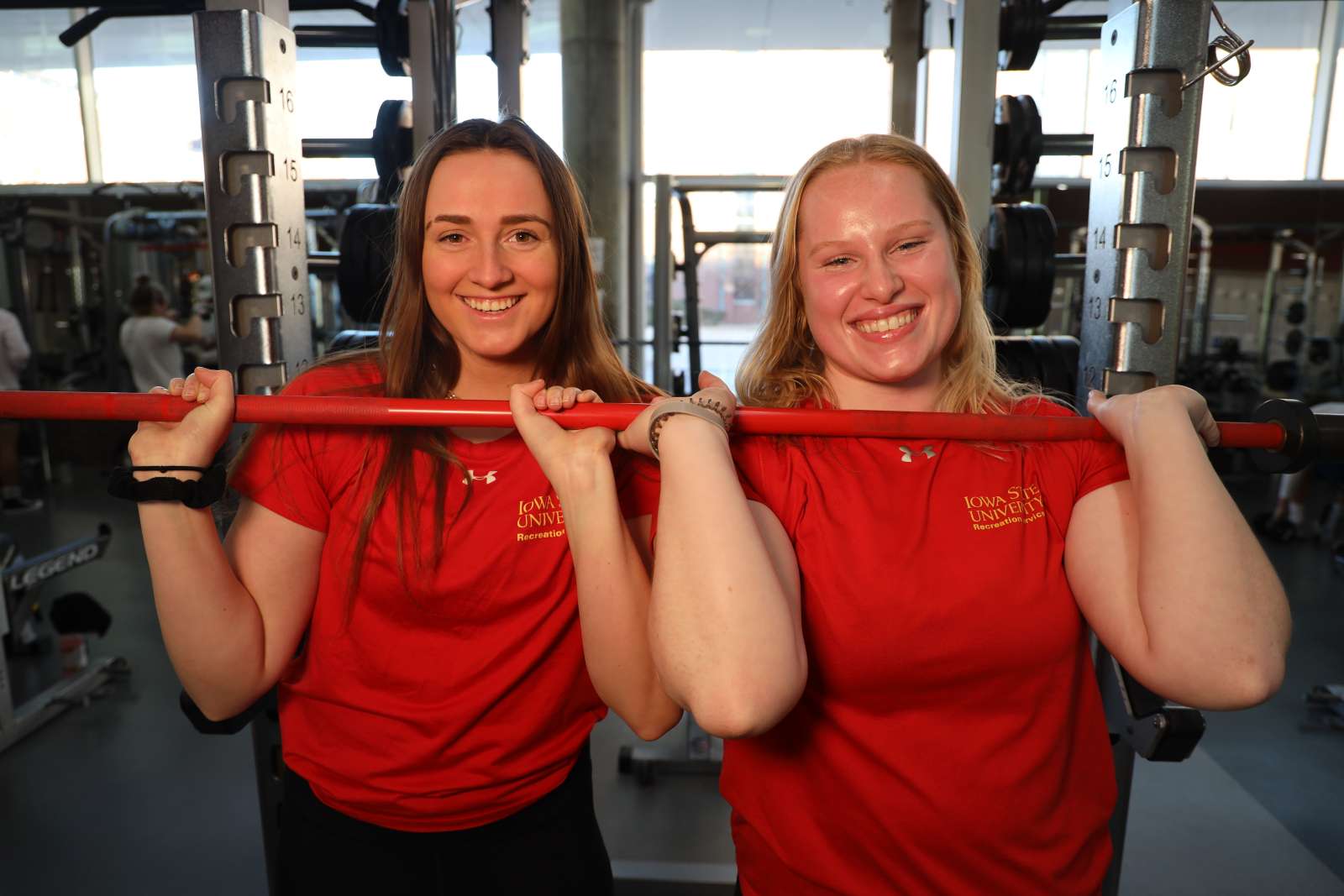 Two fitness instructors smiling while gripping a barbell in State Gym